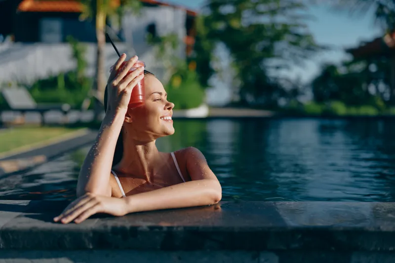 Casual poolside scene shows a joyful woman relaxed by the water, resting her elbow on the pool edge, smiling softly in a swimsuit, enjoying a carefree summer moment under sunny skies and lush