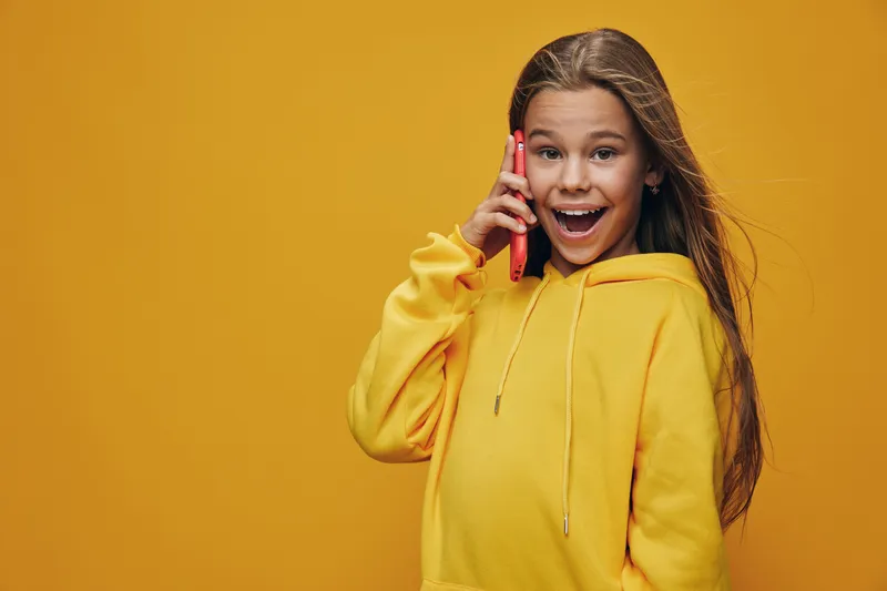 Girl in yellow hoodie smiling and talking on mobile phone with surprised happy expression against orange background. Child communication and technology concept with casual outfit.