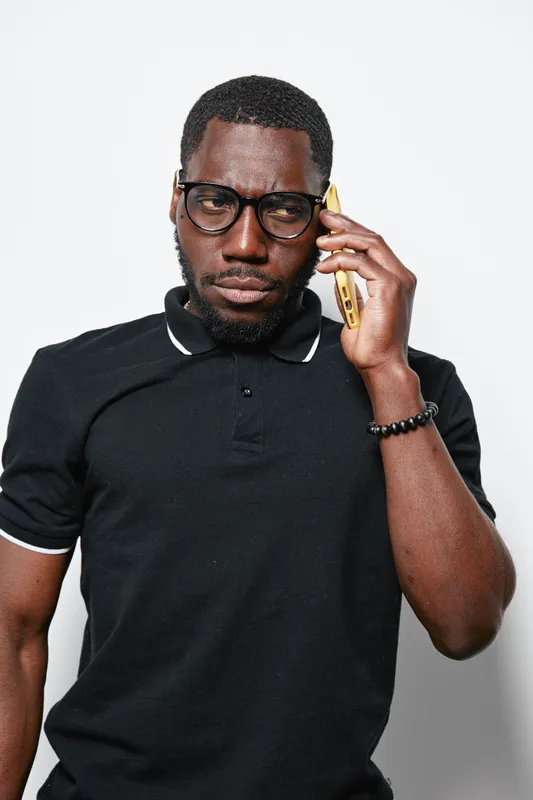 Black man with dark skin wearing glasses and black polo shirt talking on smartphone with serious expression. Adult African ethnicity person in casual outfit using mobile phone indoors.