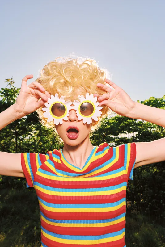 Smiling woman with curly blonde hair wearing a striped colorful shirt outdoors on a sunny day, playful retro vibe with flower glasses and vibrant mood.