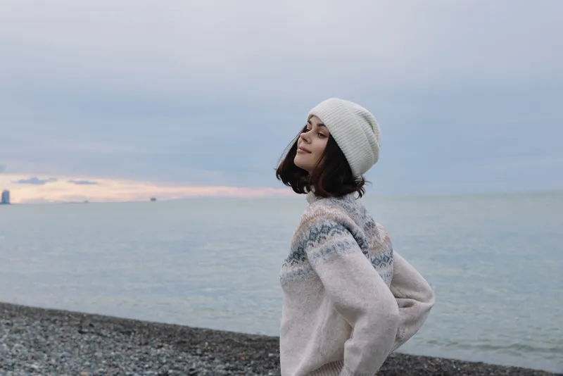 Woman on a pebble beach wearing a wool sweater and beanie, facing the sea with a calm, serene expression. Winter coastal portrait with soft light and horizon.