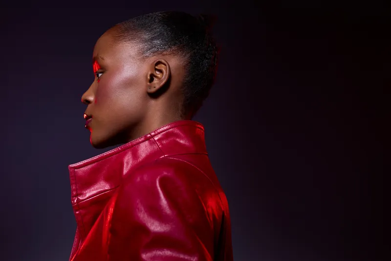 Profile portrait of a dark skinned African woman wearing a red leather jacket against a dark background. Concept of fashion, beauty, confidence, and elegant style in studio lighting.