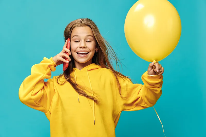 Happy girl in yellow hoodie holding balloon and talking on phone. Smiling child with closed eyes expressing joy and playfulness on blue background for fun and communication.