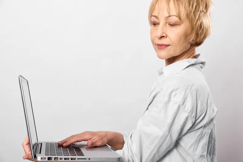 Senior woman working on laptop, hands typing on keyboard, calm expression, bright studio office light, remote technology concept, indoor workspace, focused workflow, copy space