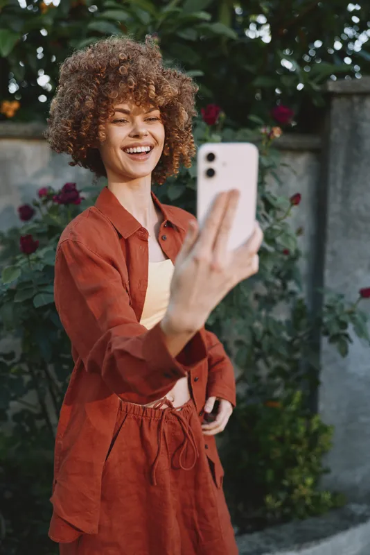 Cheerful woman outdoors, taking a selfie in a rust outfit, curly hair, bright smile, natural light, relaxed pose, lush greenery backdrop, casual lifestyle, joyful energy