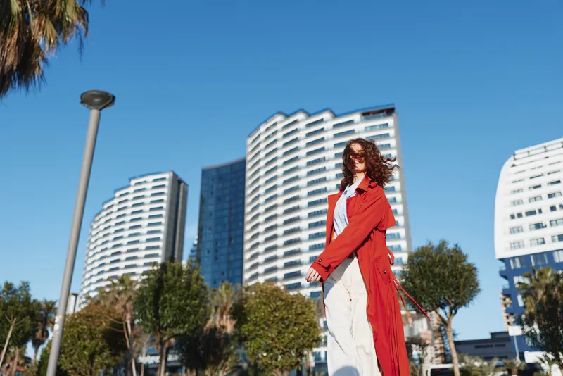 Fashionable woman walks along an urban street in a bright red coat, white dress, and sunny sky, modern buildings behind, stylish confident mood, outdoor city scene