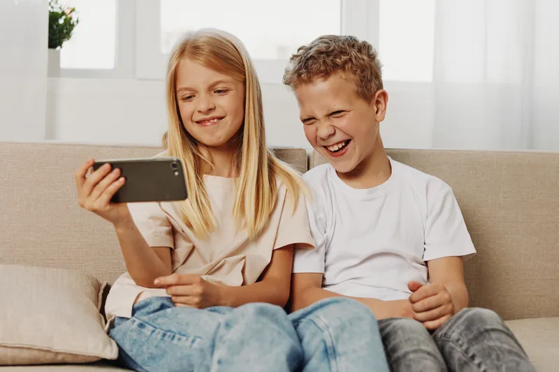 Two happy children sitting on sofa taking selfie with smartphone, smiling and laughing in bright living room. Concept of childhood, friendship, and digital technology.