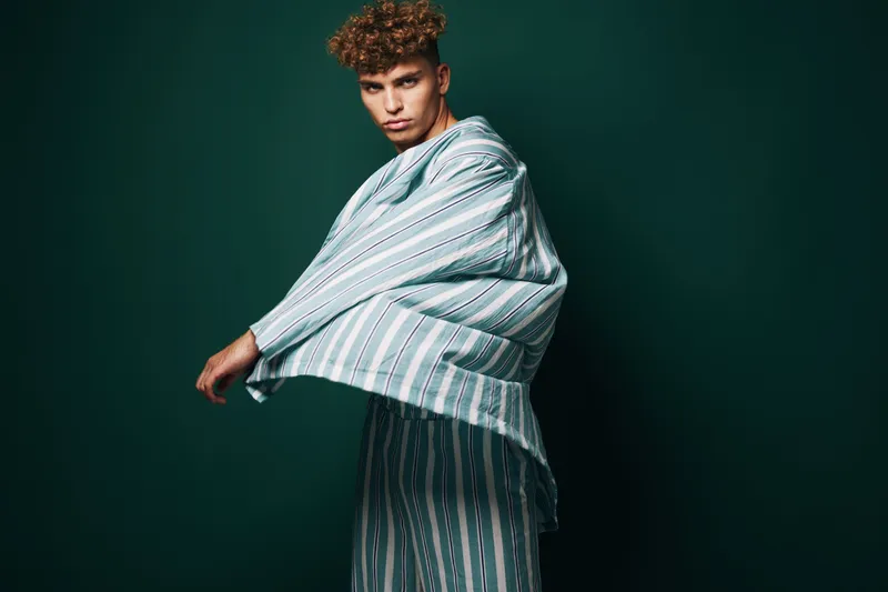 Young man with curly hair wearing striped oversized shirt and pants posing against dark green background in studio for fashion and style concept.