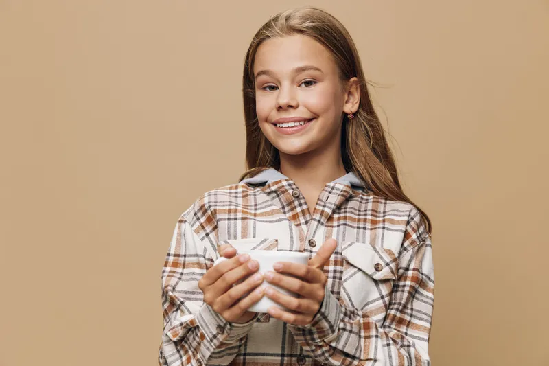 Girl smiling happily and holding white mug with both hands wearing plaid shirt on beige background. Portrait of cheerful child enjoying warm drink in cozy outfit indoors.