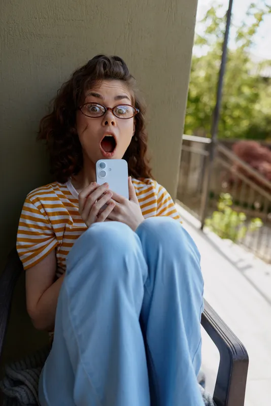 Cheerful woman indoors, surprised expression, casual outfit, striped shirt, blue pants, holding phone, sunny balcony backdrop, candid lifestyle moment, soft colors and greenery in background