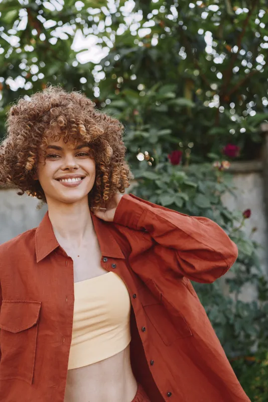Woman lifestyle portrait outdoors, smiling with curly hair, wearing a rust shirt over a pale yellow top, natural garden background, warm sunlight, relaxed casual mood, plus a soft breeze adds