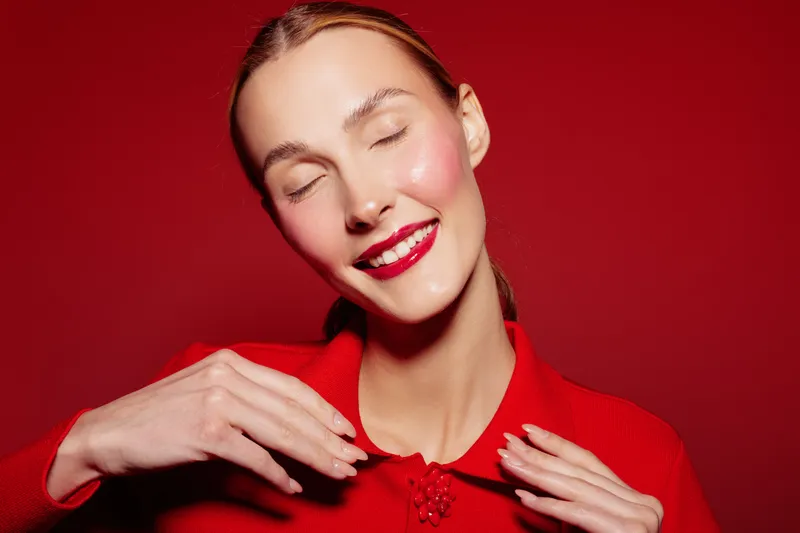 Smiling woman with closed eyes and red lipstick wears red sweater against red background. Concept of happiness, beauty, fashion, and vibrant color contrast in studio portrait.