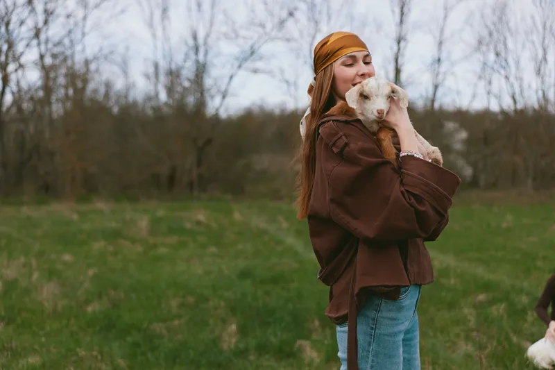 Lifestyle scene shows a smiling woman holding a goat kid outdoors, wearing a brown jacket and jeans. Warm, wholesome moment highlighting care, friendship, and simple countryside living.