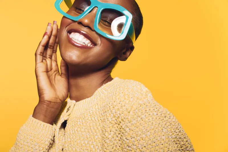 Happy African woman with dark skin wearing large blue glasses and a yellow knitted sweater smiles cheerfully on yellow background. Concept of joy, fashion, and positive mood.