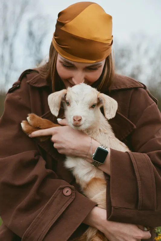 A woman in a brown coat and orange headscarf holds a goat kid in a chilly outdoor setting, expressing tenderness, care, and rustic lifestyle with farm animals.