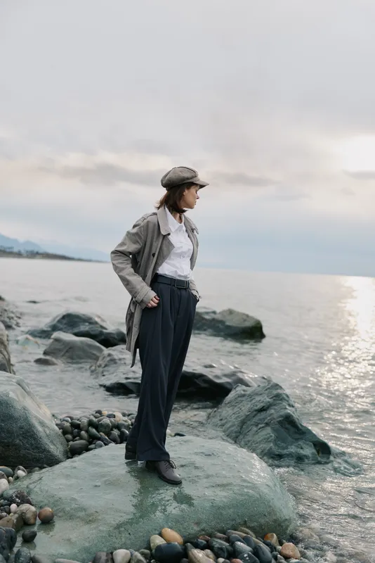 Young woman in stylish coat and hat stands on rocky shore looking at calm sea. Overcast sky and soft natural light create peaceful mood. Concept of fashion, nature, and contemplation.