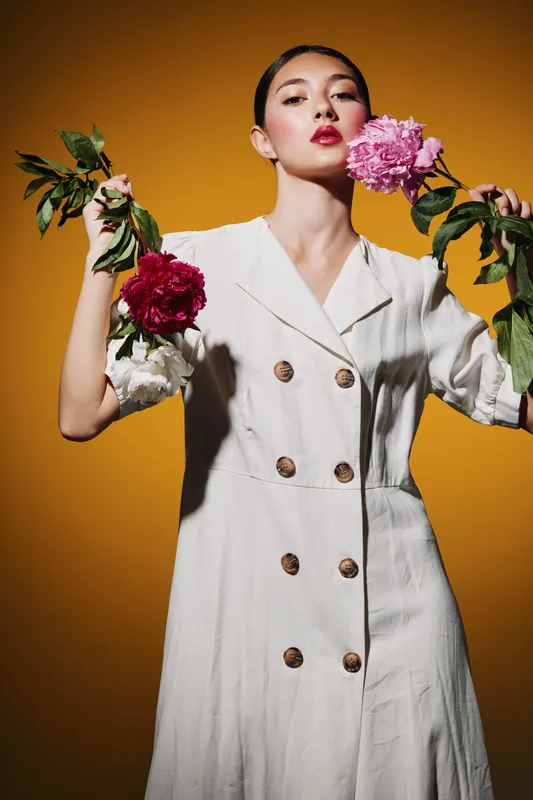 Fashion creative portrait of a young woman wearing a white dress holding two flowers. She poses against a bright orange background with confident expression. Concept for beauty and style.
