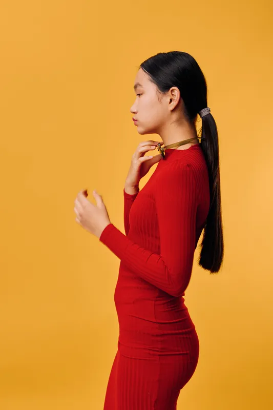 Young woman with long black hair in a red fitted dress standing sideways against yellow background. Elegant pose, ponytail hairstyle, fashion and beauty concept with colorful studio lighting.