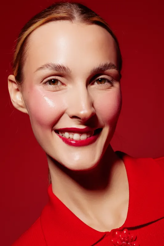 Smiling woman with red lipstick and glossy skin wears red blouse against red background. Close-up portrait with natural makeup and bright studio lighting. Concept of beauty and fashion.