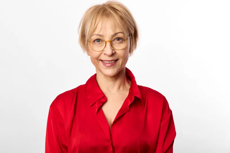 Smiling senior woman portrait in studio, wearing red blouse and glasses, neutral background, head and shoulders framing, gentle expression, calm mood, even soft lighting