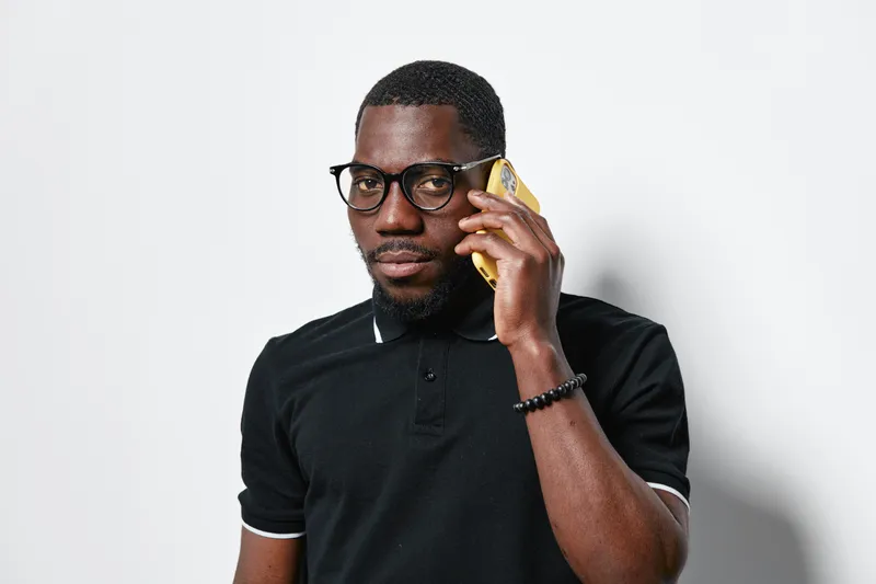 African American man with dark skin wearing glasses and black polo shirt talking on smartphone with serious expression on white background in studio.