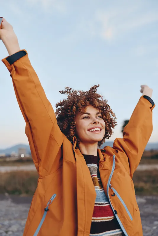 Happy woman lifestyle outdoors in autumn field, wearing orange coat, curly hair, raises arms and smiles to bright sky, casual portrait, warm natural light
