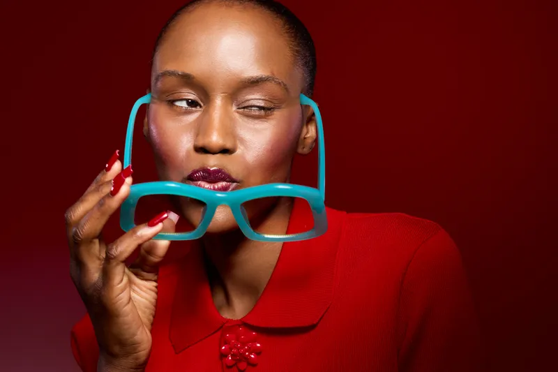 Close-up of a stylish black woman with dark skin holding turquoise glasses near her face. She wears a red outfit and poses against a red background. Concept of fashion and beauty.