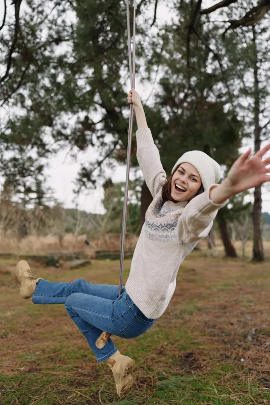 Girl on a swing outdoors, smiling and carefree, wearing a knit sweater, jeans, boots and a beanie in a park setting with trees and autumn light.