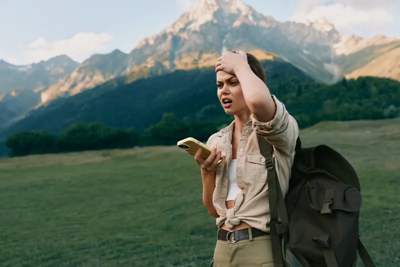 A young woman with a backpack stands in a mountain field holding a smartphone and looks confused while touching her head. Concept of outdoor lifestyle and navigation challenges.