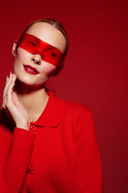Stylish woman in red sweater and red translucent sunglasses poses against red background. Fashion portrait with glossy lips and smooth skin. Concept of modern style and bold color.