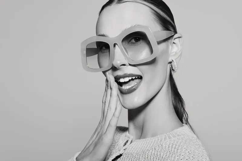 Young caucasian woman with fair skin wearing large sunglasses and earrings shows surprised expression. Studio portrait in black and white with casual sweater and slicked hair style.