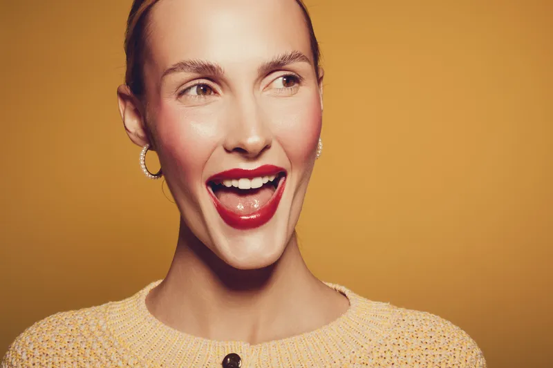 Happy woman with red lipstick and gold earrings smiles broadly against warm yellow background. Close-up portrait with natural skin and joyful expression. Concept of beauty and happiness.