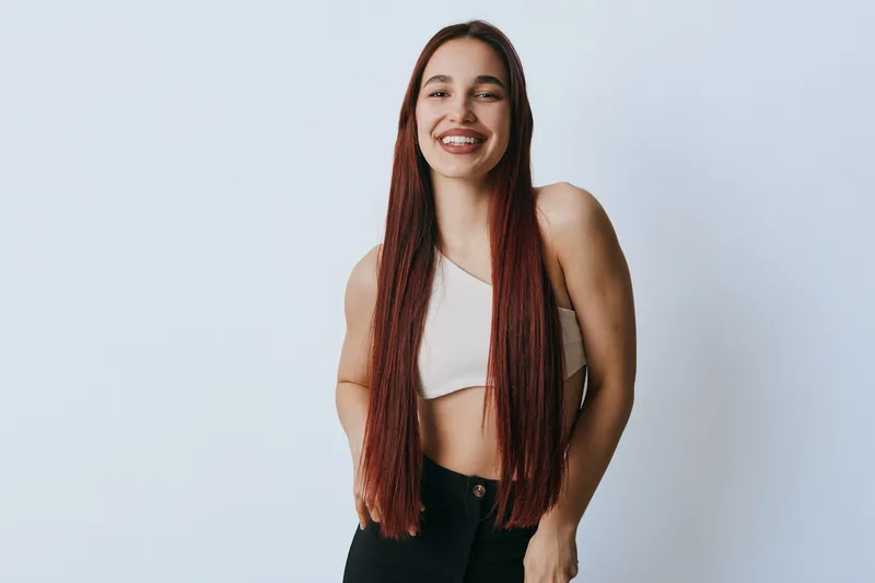 Young woman with long straight hair smiling and posing in casual outfit with white crop top and black pants on plain light background in studio. Female model with confident posture.