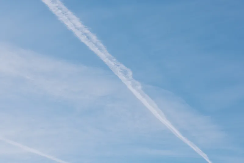 Blue sky with a diagonal white contrail crossing the frame, wispy clouds and expansive atmosphere, conveying openness, travel inspiration and quiet aviation mood in daylight.