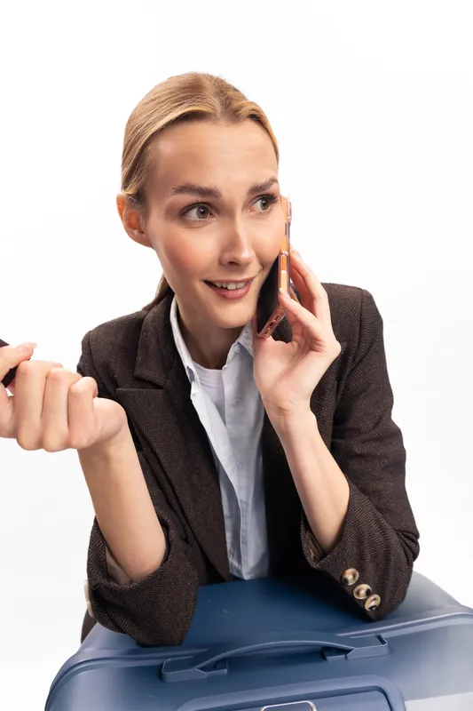 Young woman in business attire talking on smartphone while resting on blue suitcase. Concept for travel, business trip, communication, and professional lifestyle with white background.
