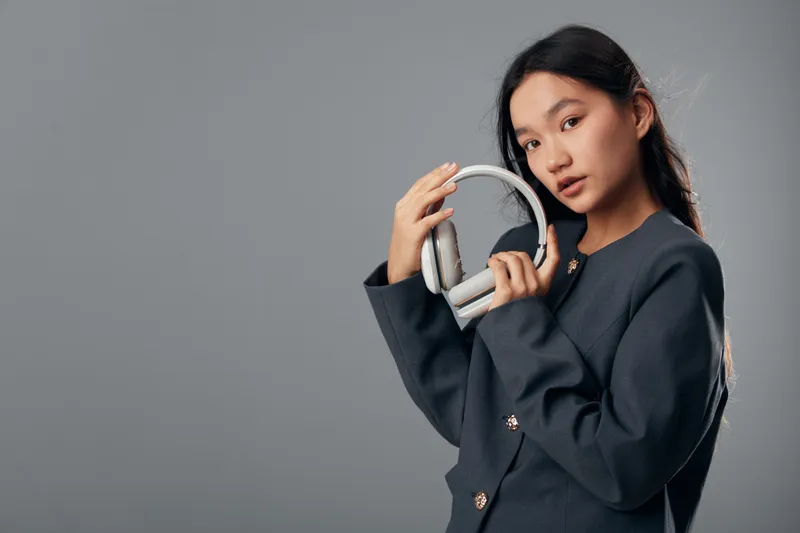 Young woman with long hair holding white headphones wearing dark blazer against gray background. Portrait of female with modern audio device for music or technology lifestyle concept.