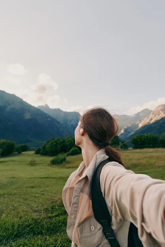 Woman in outdoor landscape with backpack stands in green field facing mountains, taking a selfie to capture travel and nature lifestyle and a cheerful mood enhances the moment.