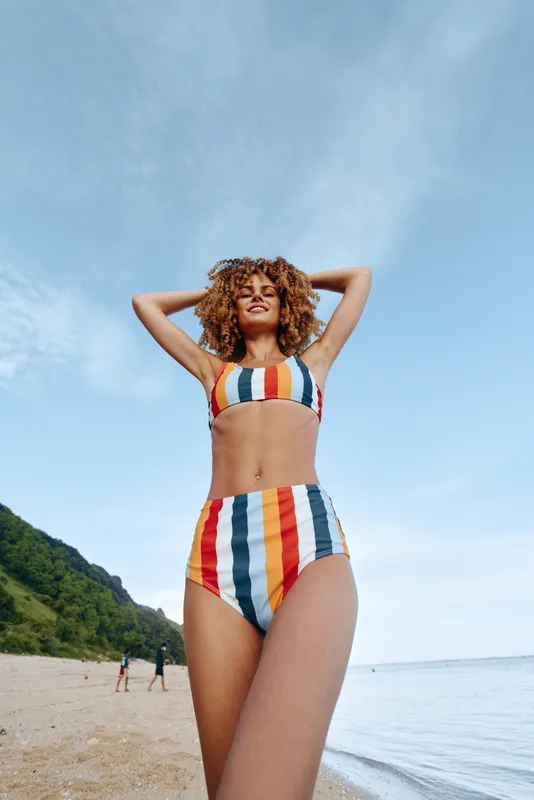 Woman in bikini on beach by ocean in summer vacation, hands behind head, curly hair, striped swimsuit, calm smile, clear blue sky, green hills, natural daylight, travel lifestyle