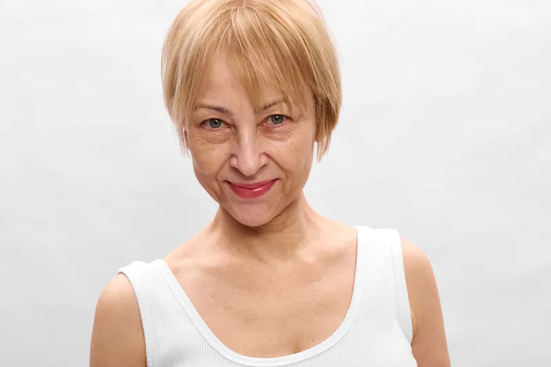 Elderly woman, portrait, studio headshot, smile, short blond hair, wearing white sleeveless top, neutral background, calm friendly expression, soft natural light, closeup face
