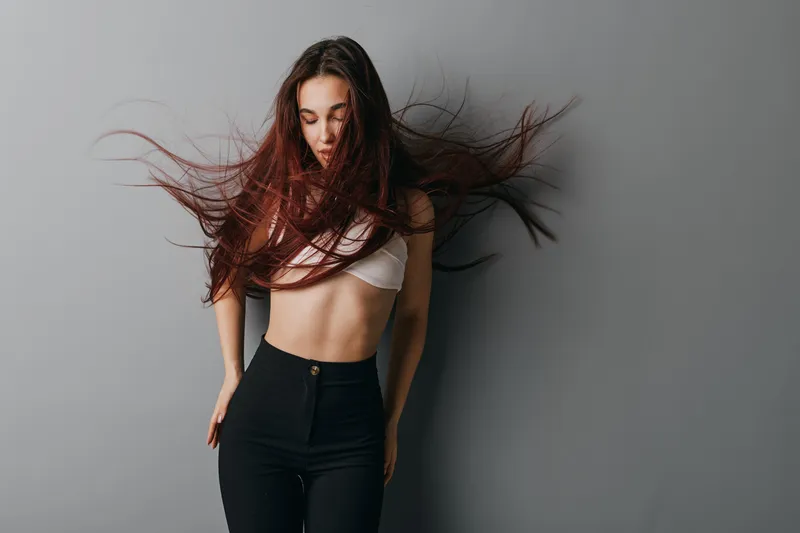 Young woman with long flowing hair in motion wearing white crop top and black pants standing against gray background with closed eyes and relaxed expression in studio.