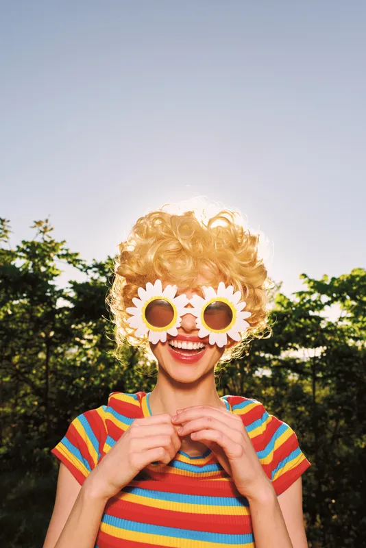 Smiling woman with curly hair wearing flower sunglasses in a sunny park. Retro film color mood, outdoor portrait with a vibrant striped shirt, casual lifestyle moment.