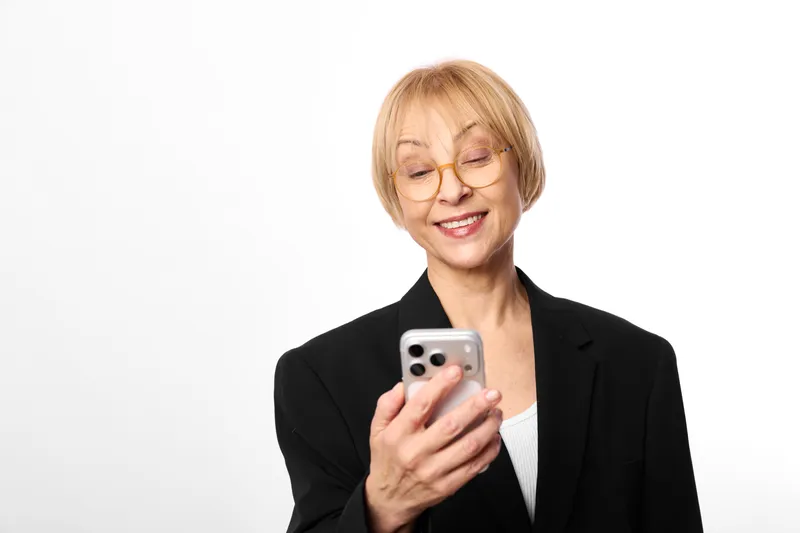 Smiling senior woman holding a smartphone for communication, portrait on a white studio background, black blazer and glasses, modern technology concept and call.