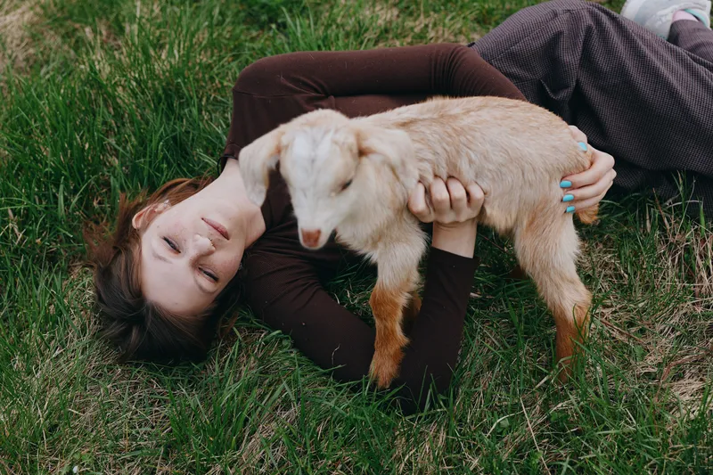 Relaxed woman lying on grass cuddling a young goat kid in a sunny outdoor setting, showcasing rural lifestyle, animal companionship, and gentle bonding. Soft light and calm mood. Warm.