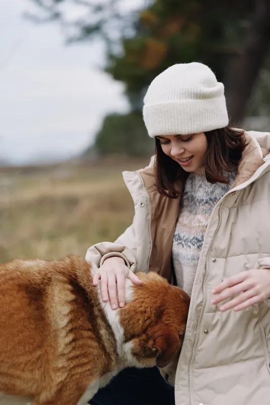 Woman in a warm coat and beanie pets a red panda outdoors among a grassy field with blurred trees showcasing gentle animal interaction and serene nature moments