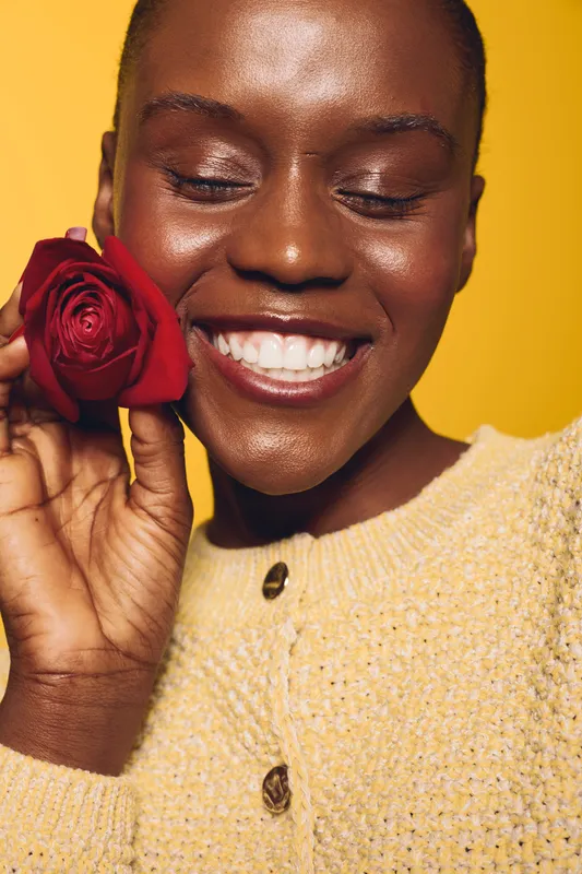 Happy African woman with dark skin holds red rose near face smiling with closed eyes. Yellow background and cozy beige sweater create warm, positive mood. Concept of beauty and joy.