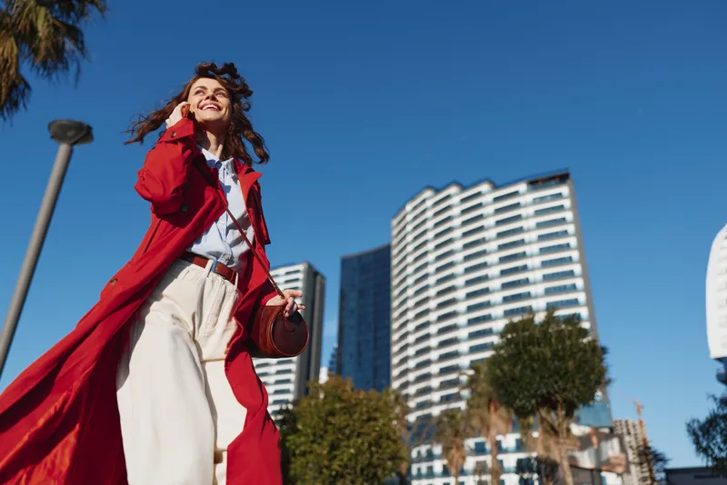 Smiling woman walks along a sunny city street in a long red coat, urban skyline behind, stylish fashion moment captured outdoors with breeze and confidence.