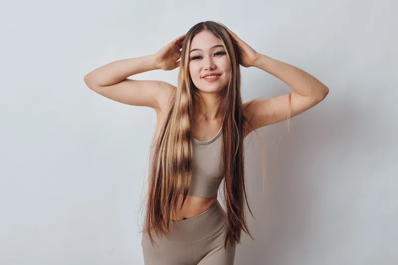 Young woman with long braided hair posing with hands on head wearing beige tank top and leggings against white background. Beauty, confidence and fashion concept in studio.