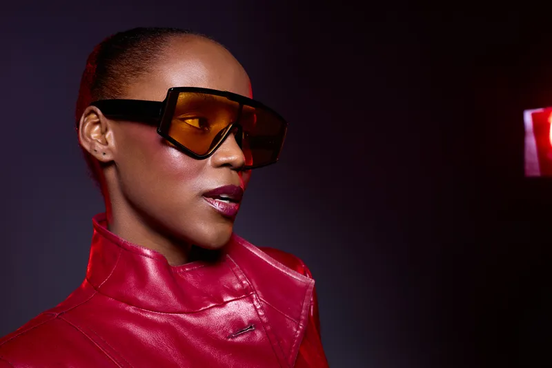 Stylish African woman with dark skin wearing oversized orange sunglasses and a red leather jacket poses confidently in a studio with dramatic lighting. Fashion and attitude concept.