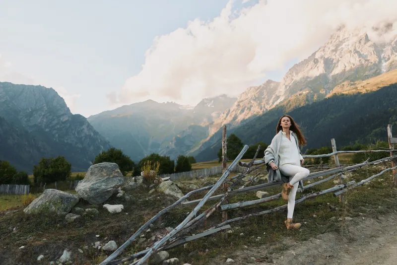 Woman in white casual attire stands beside a rustic wooden fence in a mountain valley, capturing a lifestyle moment with nature, travel, and calm wellness vibes. Soft light.