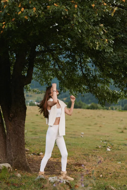 Young woman in white clothes stands under a large tree in a grassy field enjoying a peaceful moment in nature during daytime.